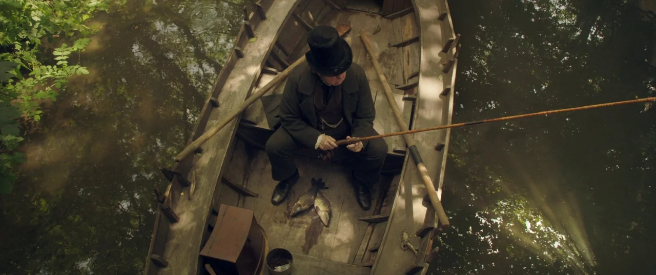 Mr. Turner (2014) movie still: fullbody — A wooden rowboat floats on a shaded river surrounded by dense overhanging green trees. A man in a…