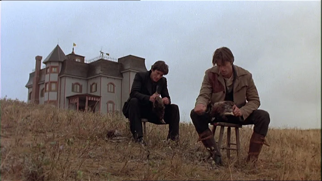 Days Of Heaven (1978) movie still: wide — A large, multi-story country mansion sits on a grassy hill under an overcast sky. Two men sit on small…