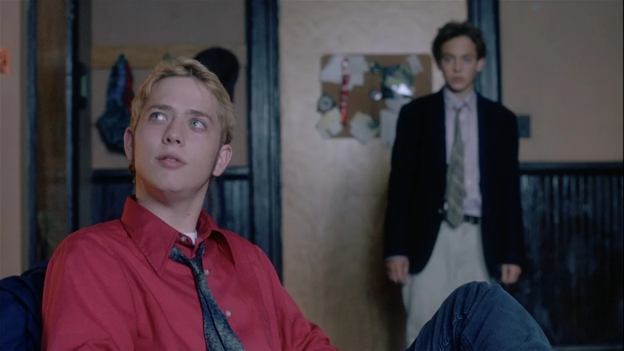 Boys (2021) movie still: medium — An interior school setting with dark wooden trim and a bulletin board behind occupants. A young man sits in the…