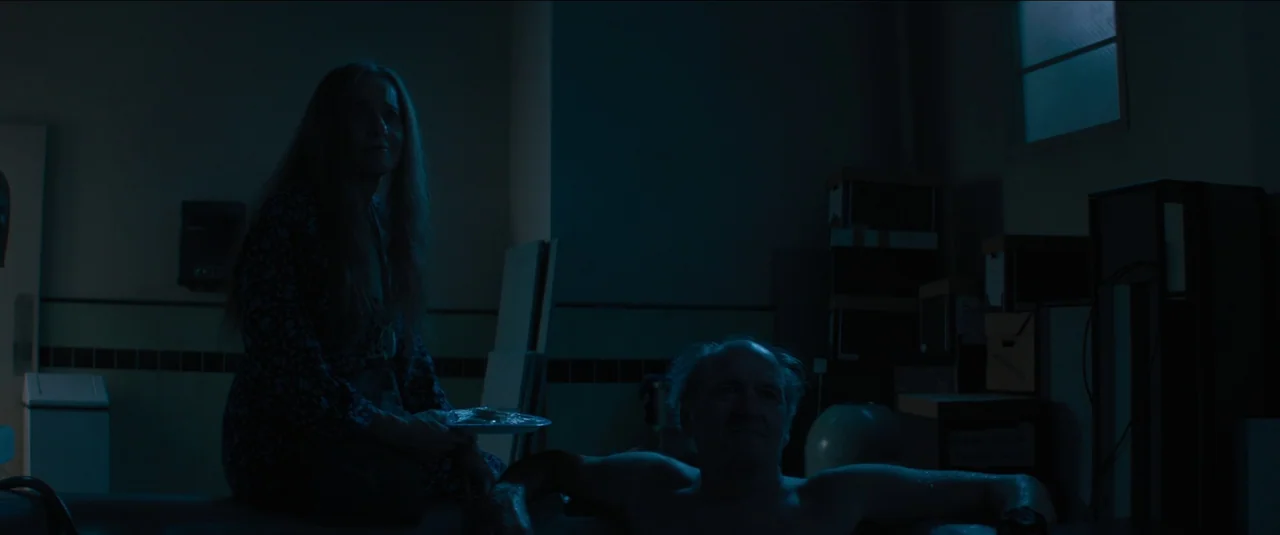 Kajillionaire (2020) movie still: medium — A dimly lit basement features cluttered cardboard boxes and stacks of wood planks. A man and woman sit…