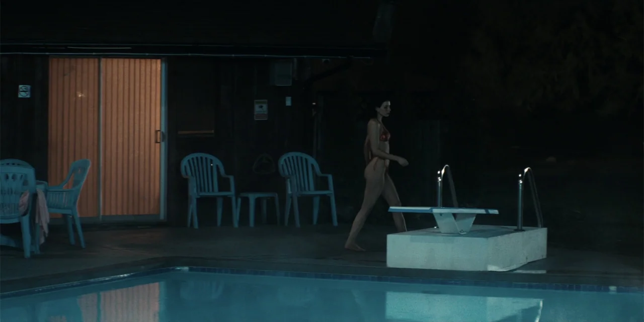 The Monkey (2025) movie still: fullbody — A night view of a residential backyard with a rectangular swimming pool and patio furniture. A woman in a…