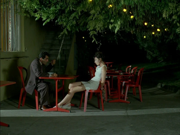 Helas Pour Moi (1993) movie still: fullbody — An outdoor patio at night features red chairs and tables under dark tree foliage. A man and a woman…
