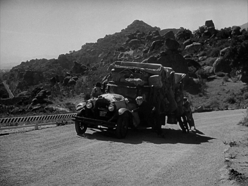 The Grapes of Wrath (1940) movie still: wide — A rugged mountain landscape with rocky hills borders a winding paved road. A family pushes a heavily…