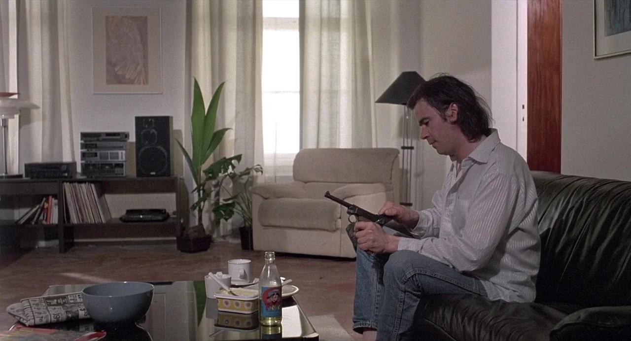 Ariel (1988) movie still: medium — A living room features a black leather sofa, a glass coffee table, and a stereo system. A man sits on the sofa…