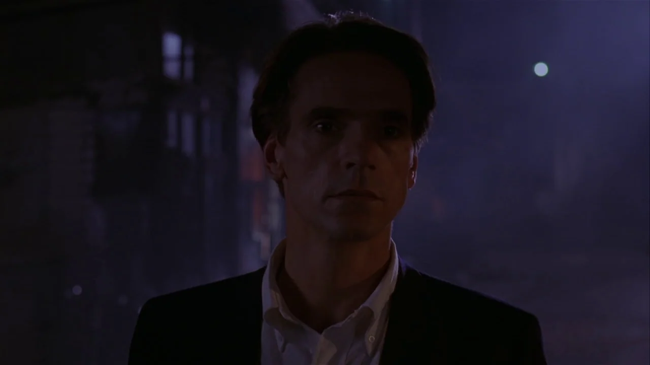 M. Butterfly (1993) movie still: medium — A man stands on a dark, dimly lit city street at night. He is centered in the frame with an obscured urban…