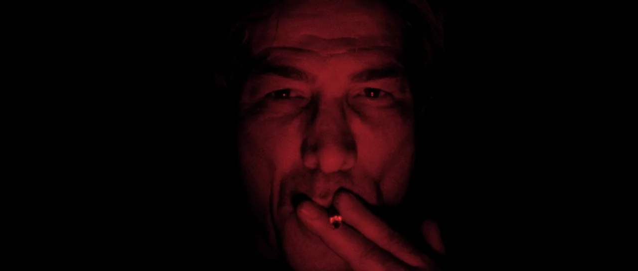 Let The Corpses Tan (2017) movie still: closeup — A dark, featureless environment surrounds a man's face lit by an intense red source. The subject…