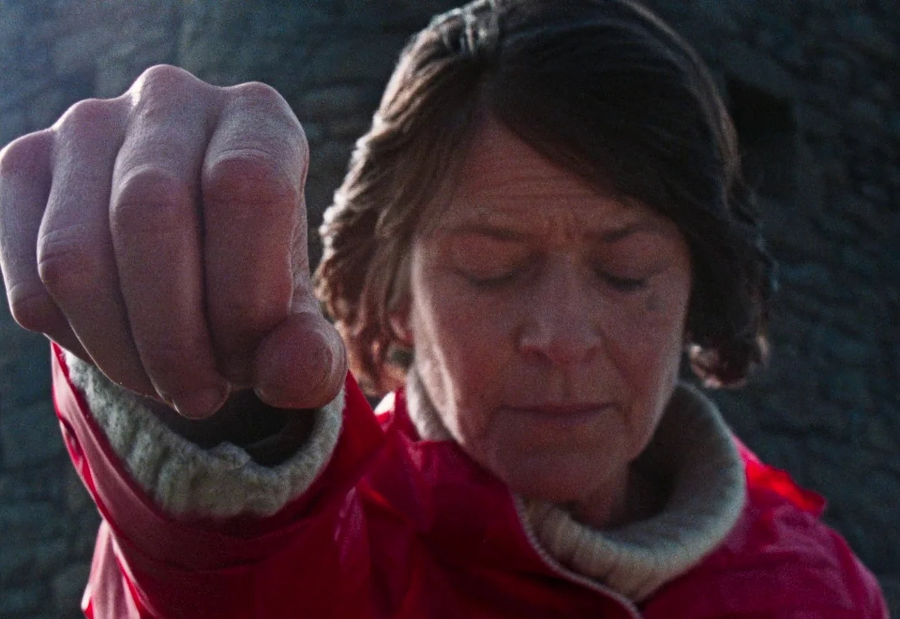 Enys Men (2022) movie still: closeup — A woman stands before a dark, textured stone wall in daylight. She holds one hand extended forward toward the…