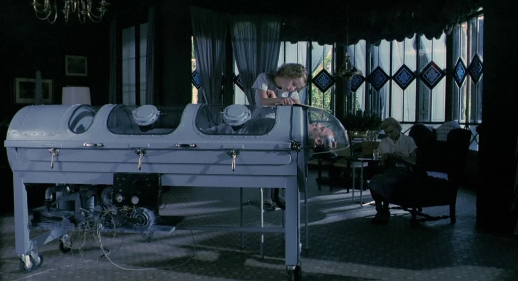 In a Glass Cage (1986) movie still: wide — A dark, ornate room with floor-to-ceiling windows features a large vintage medical iron lung. A young…