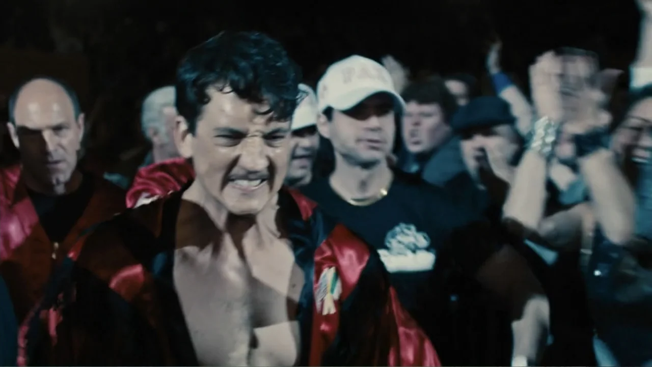 Bleed for This (2016) movie still: medium — A crowded, dimly lit industrial hallway lined with people watching a fighter pass by. The athlete moves…