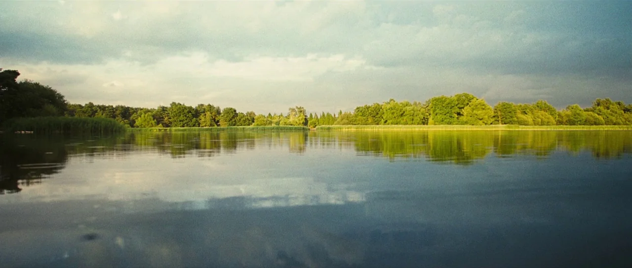 Eden Lake (2008) movie still: establishing — A calm lake stretches across the horizon under a cloudy daytime sky. Dense green trees and tall reeds…