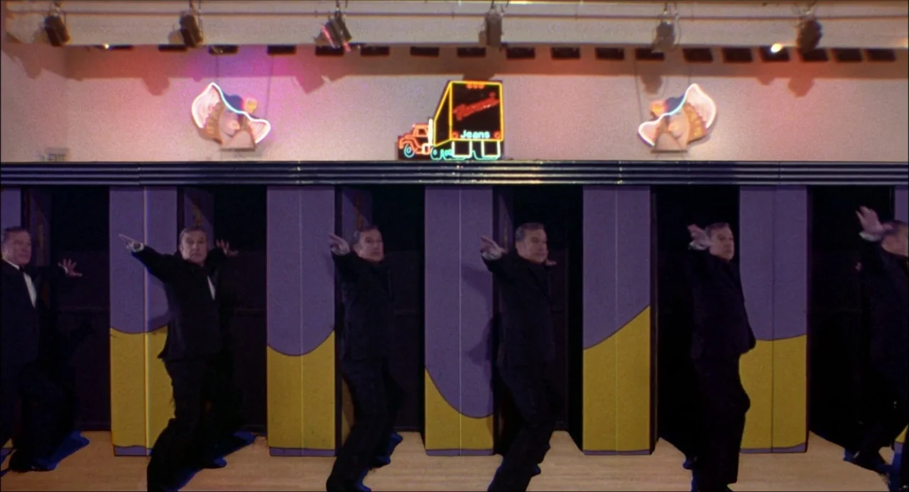 Xanadu (1980) movie still: fullbody — A performance space features purple and yellow partitions beneath neon signage on a wall. A man in a black…