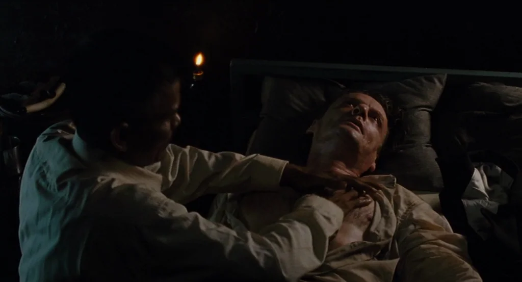 Almayer’s Folly (2011) movie still: medium — A dark bedroom with minimal light illuminating the bed and two men. One man reclines on the mattress…