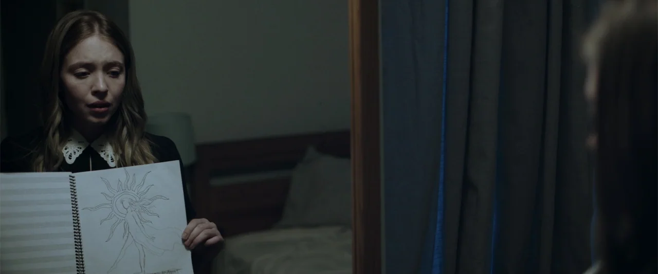 Nocturne (2020) movie still: medium — A dimly lit room with a bed visible in the blurred background. A young woman in a lace-collared top holds a…