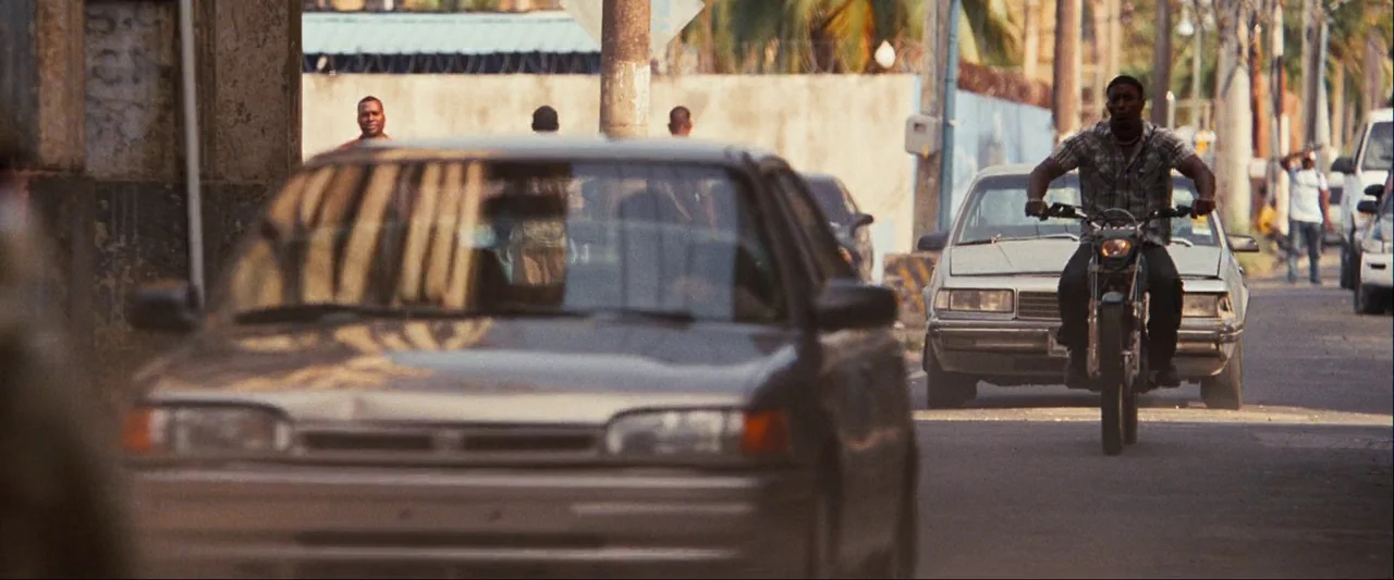 Quantum of Solace (2008) movie still: wide — An urban street features multiple motor vehicles and pedestrians walking in the background. A man rides…