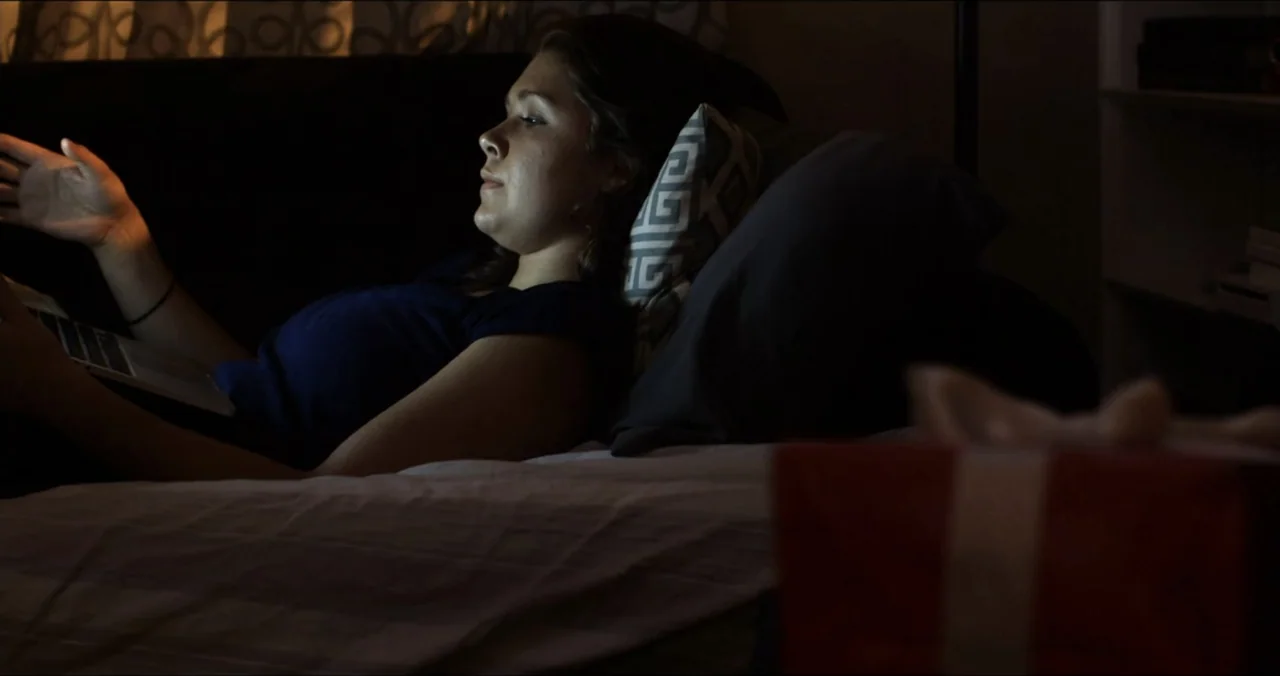 Faith, Love & Chocolate (2018) movie still: medium — A dark bedroom with a bed featuring pillows and a wrapped gift box nearby. A woman reclines…