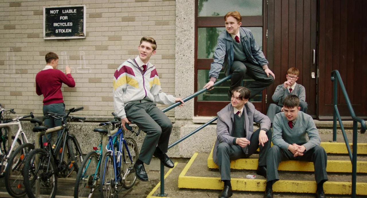 Dating Amber movie still: medium — A group of six teenage boys in school uniforms congregate on and around steps outside a brick building. One boy…
