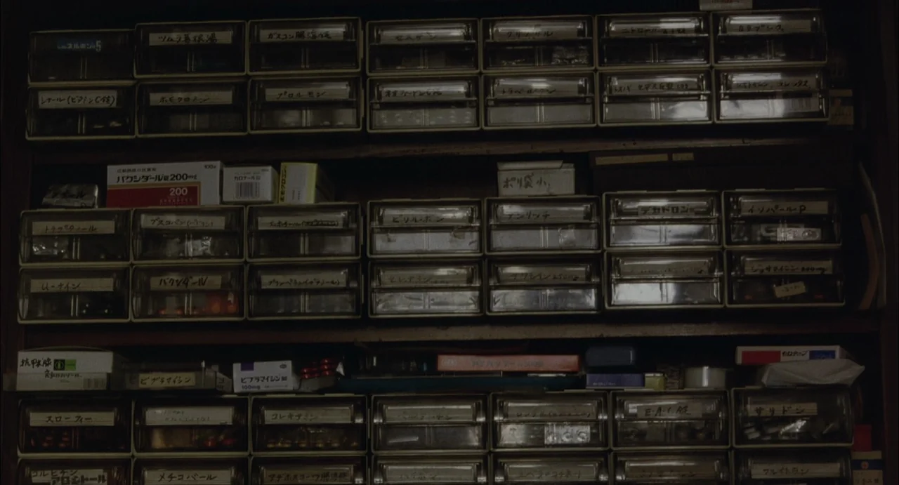 Still Walking (2008) movie still: closeup — A dense collection of plastic medical drawers filled with labeled medication boxes and pills. Rows of…