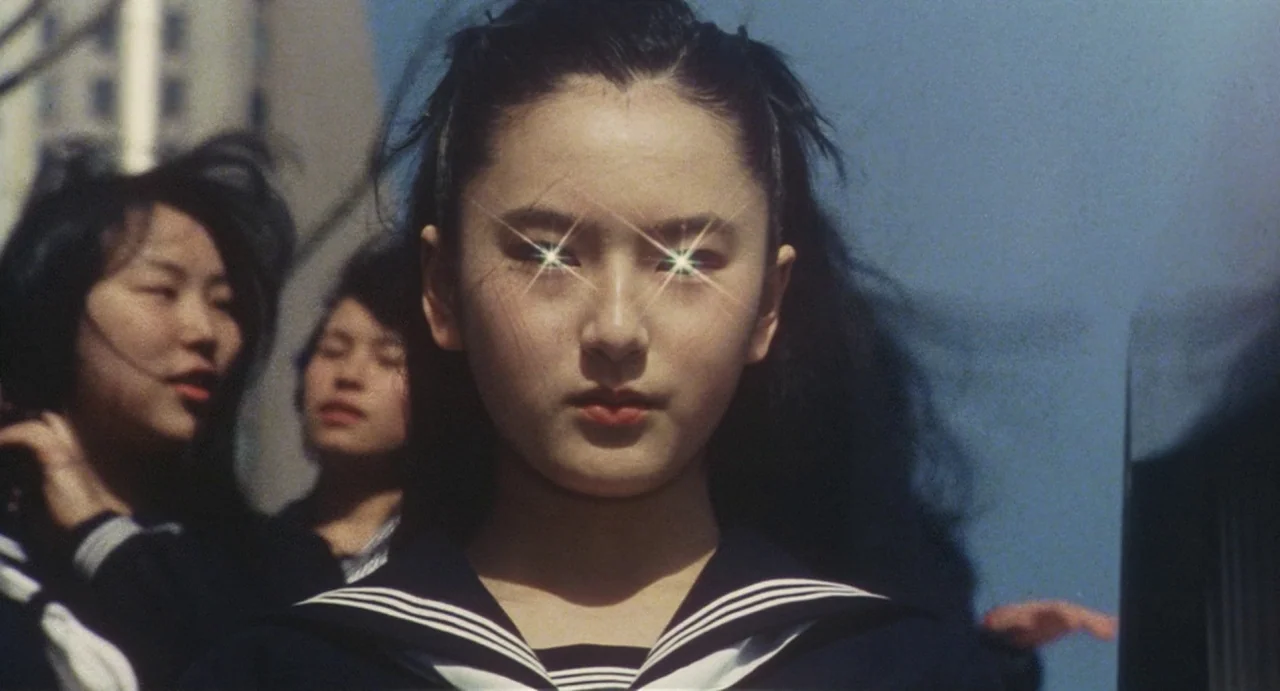 School in the Crosshairs (1981) movie still: closeup — A teenage girl stands outdoors in front of other students under a clear blue sky. She wears a…