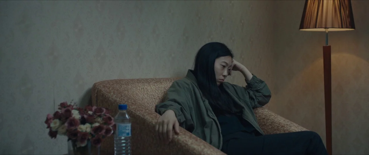 The Farewell (2019) movie still: medium — A dimly lit hotel room features patterned wallpaper and a floor lamp. A woman sits in a textured armchair…