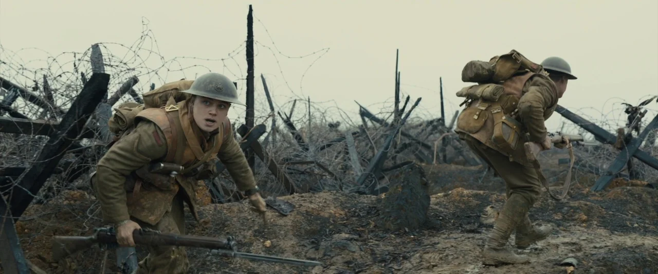 1917 movie still: fullbody — A desolate battlefield features scorched earth and tangles of jagged, weathered barbed wire fencing. Two soldiers in…