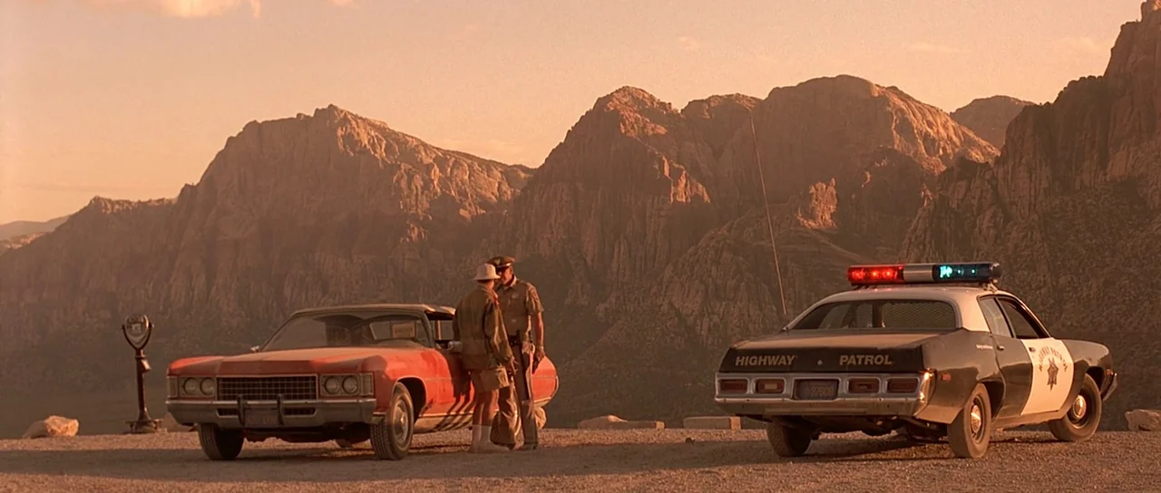 Fear and Loathing In Las Vegas (1998) movie still: wide — A desolate gravel turnout sits before a vast, craggy mountain range under orange light. An…
