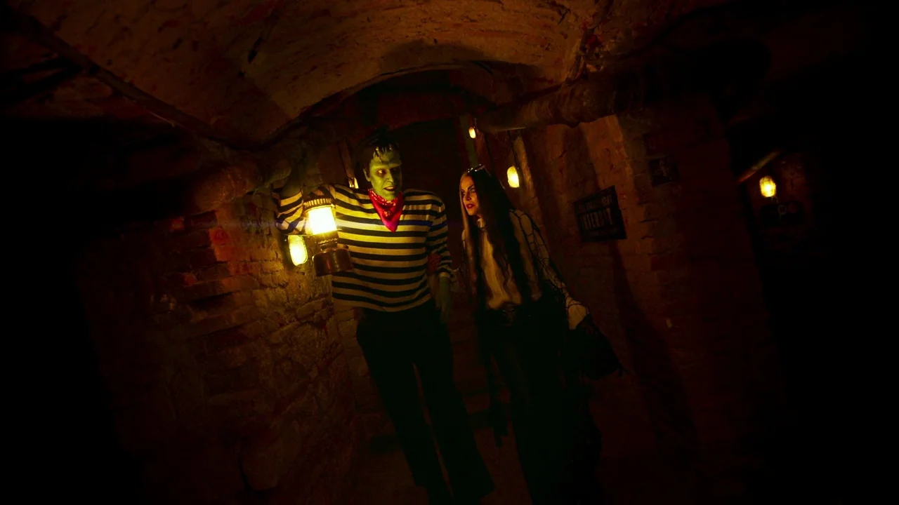 The Munsters (2022) movie still: medium — A dark brick subterranean tunnel has low vaulted ceilings and exposed piping on walls. A man with green…