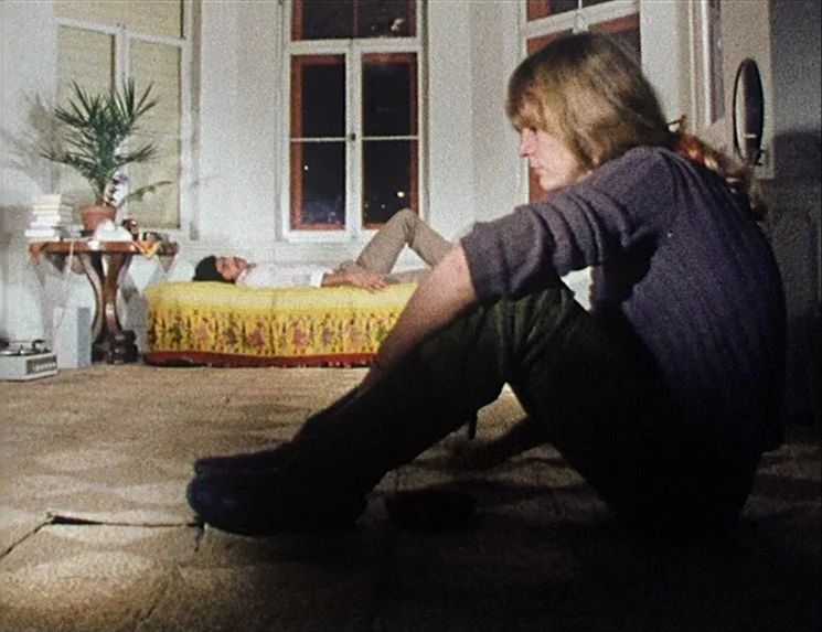 Rio Das Mortes (1971) movie still: medium — A living room has patterned carpet and a bed positioned against a bay window. A woman sits on the floor…