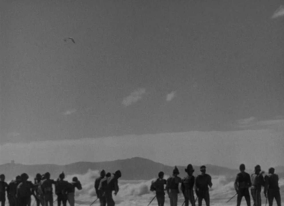 Othello (1952) movie still: wide — A wide sandy beach under an overcast sky with breaking surf in the distance. Soldiers in military uniforms stand…
