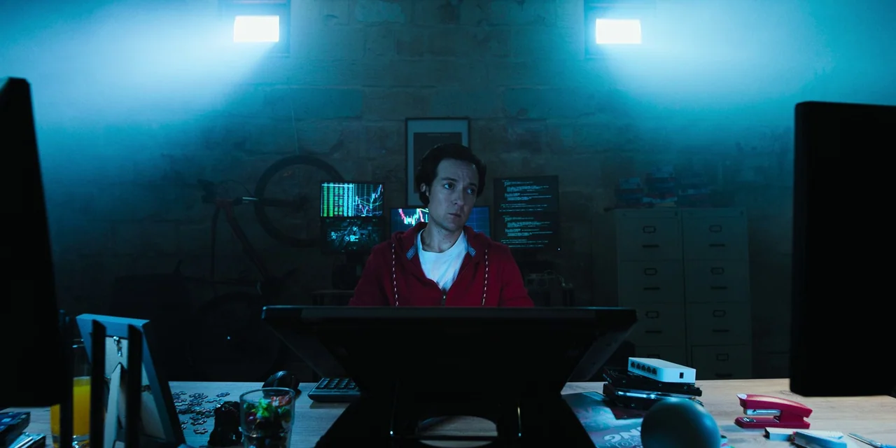 Sniper: G.R.I.T. (2023) movie still: medium — A basement room with rough stone walls features multiple monitors and computer equipment. A man sits…