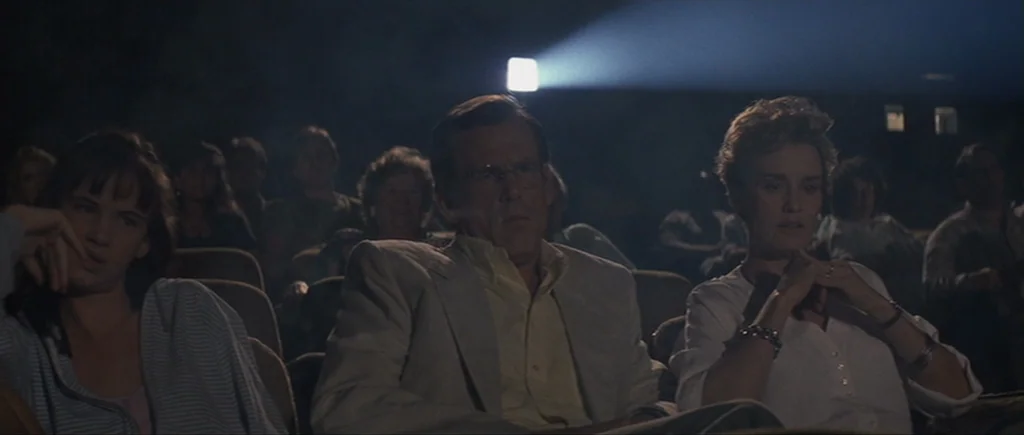 Cape Fear (1991) movie still: medium — A dimly lit movie theater filled with an audience watching a film. Three primary subjects sit in a row while…