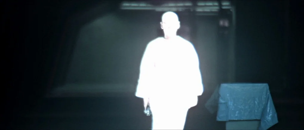 THX 1138 (1971) movie still: medium — A dimly lit, dark space contains a single brightly illuminated, overexposed figure standing centrally. Beside…