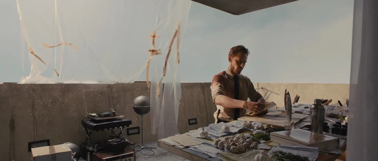 High-Rise (2015) movie still: medium — A concrete balcony features a large table scattered with garlic bulbs and loose papers. A man stands at the…