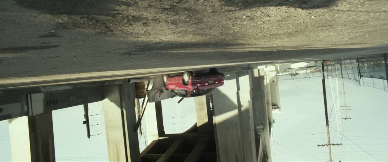 Maniac (2012) movie still: wide — A concrete overpass structure spans a wide area with large support pillars. A red passenger car is parked on the…