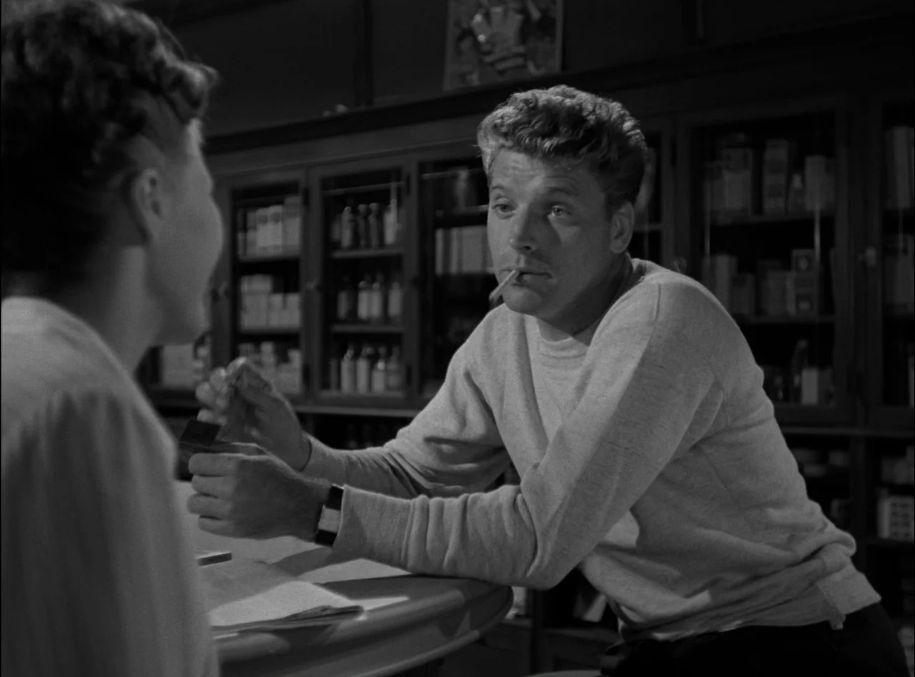 Criss Cross (1949) movie still: medium — A pharmacy interior features dark wooden shelving units filled with glass medicine bottles. A man sits at a…