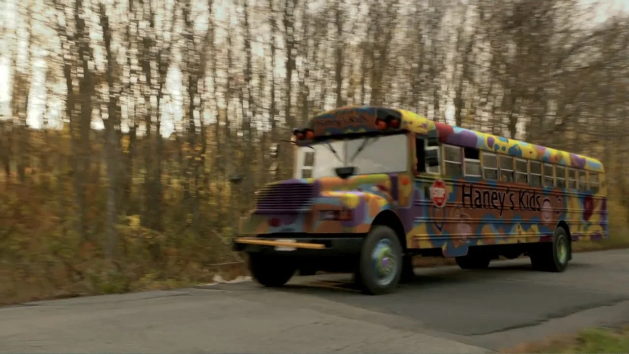 Clear History (2013) movie still: fullbody — A multicolored school bus travels along a rural road lined with bare deciduous trees. The bus features…