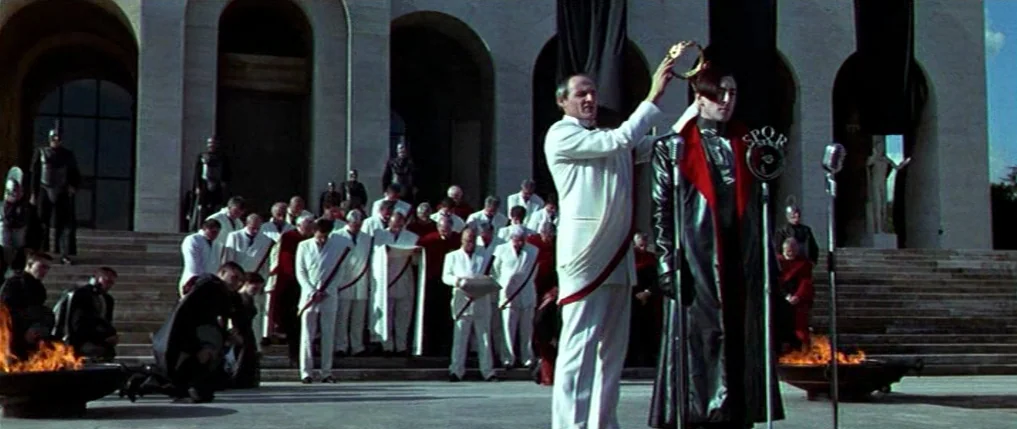 Titus (1999) movie still: wide — A stone monument with large arched doorways serves as the backdrop for a ceremony. An older man in a white toga…