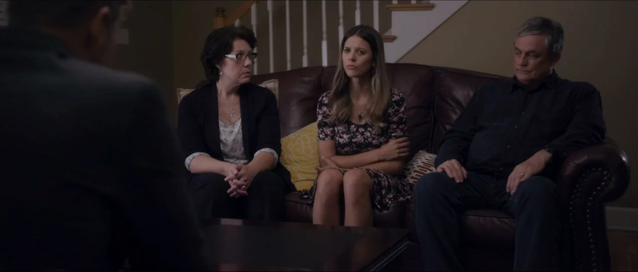 If You're Gone (2019) movie still: medium — A living room features a dark brown leather sofa against a tan wall. Three adults sit on the sofa facing…
