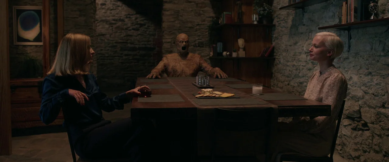 Oddity (2024) movie still: wide — A stone-walled dining room features a dark wood table with a cheese platter. Two women sit at opposite ends of the…