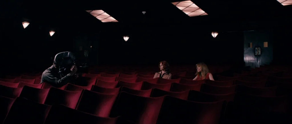 The Look of Love (2013) movie still: wide — A dark, empty movie theater features rows of red velvet seats in low light. Two women sit spaced apart…