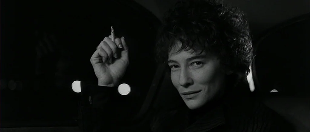 I’m Not There (2007) movie still: closeup — A person sits inside a dark vehicle at night with city lights out of focus. A hand holding a cigarette…