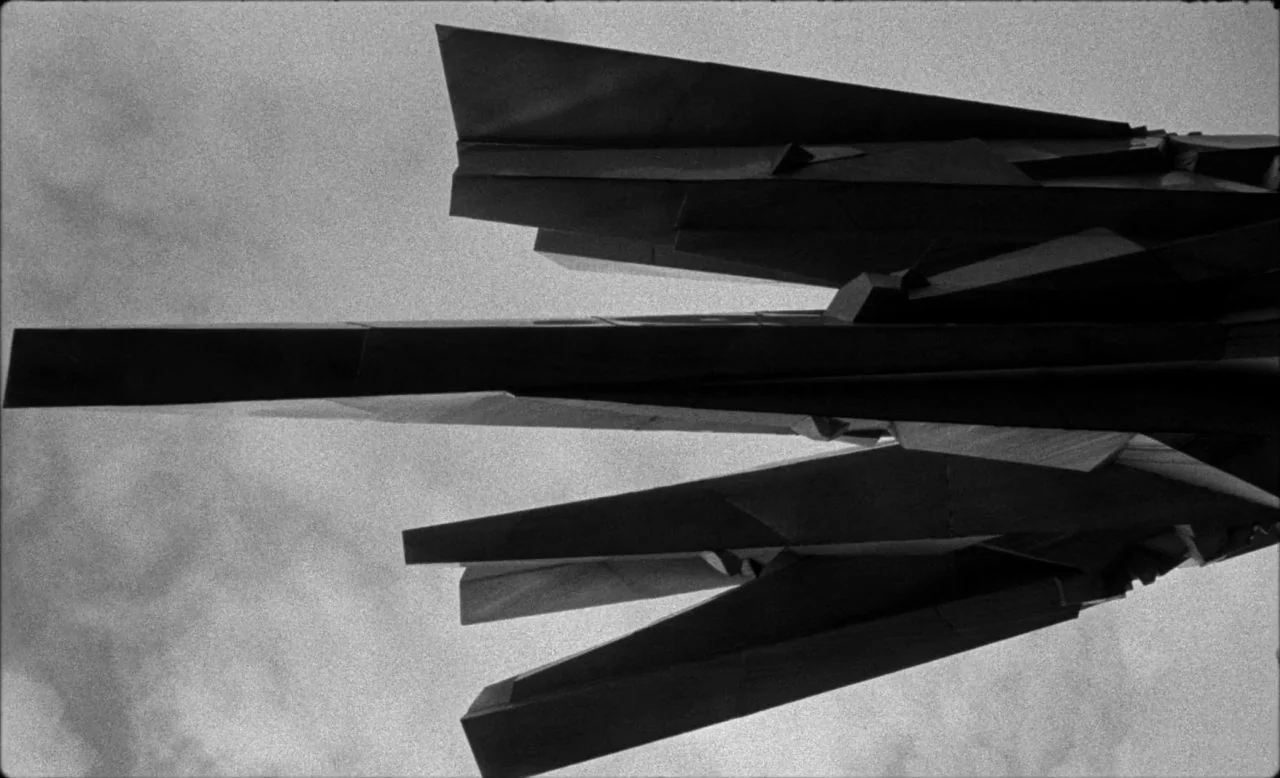 Last and First Men (2020) movie still: establishing — An angular, multi-faceted metal sculpture rises against a cloudy, bright sky. The geometric…