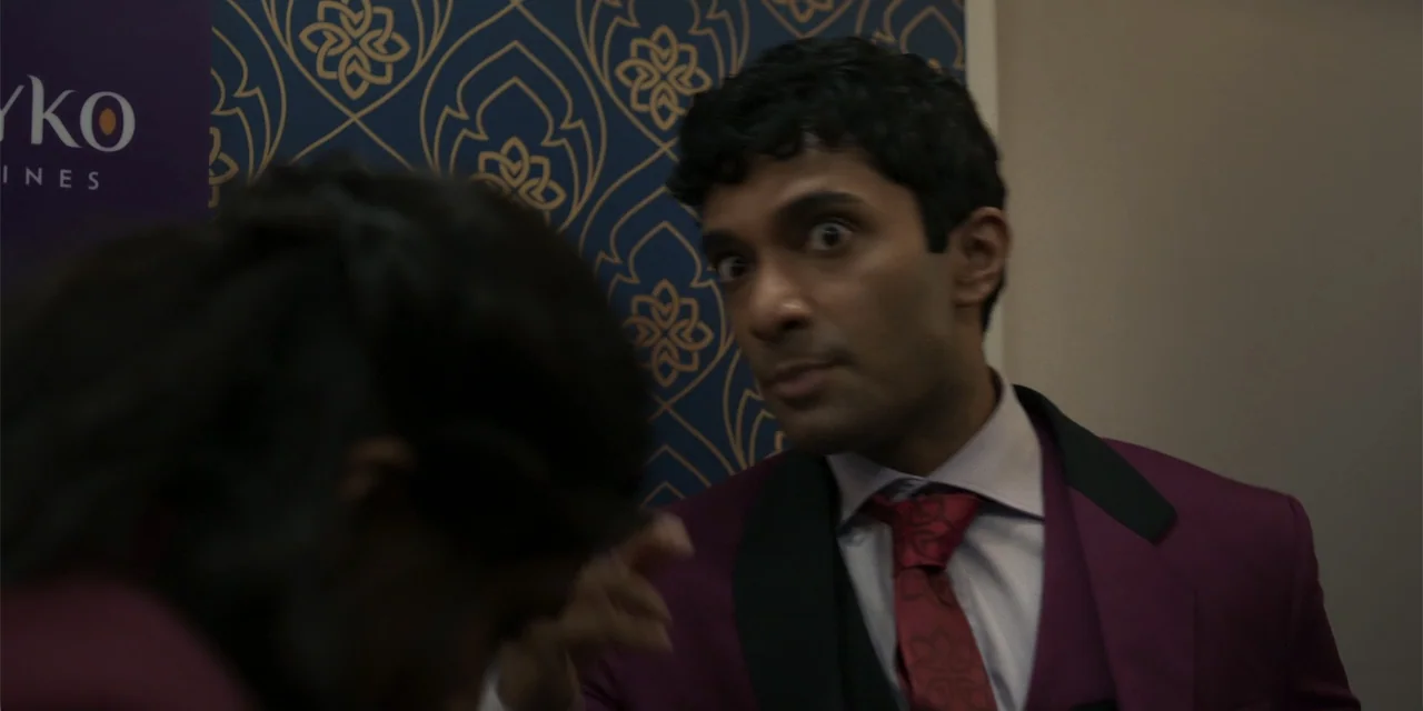 Fight or Flight (2025) movie still: medium — A man in a formal purple suit stands before a patterned blue and gold wallpaper wall. He maintains eye…