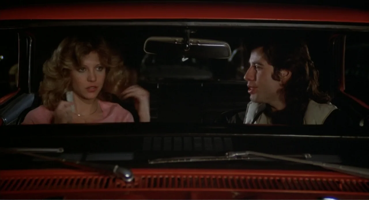 Carrie (1976) movie still: medium — A man and woman are seated inside a red vintage car at night. They are looking at each other while inside the…