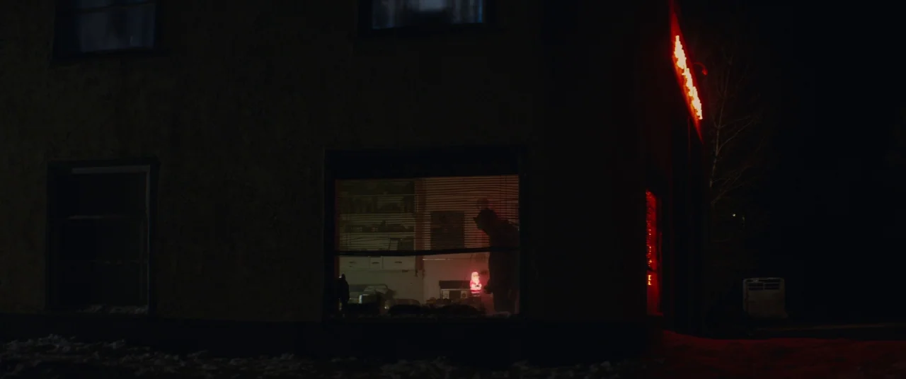 Hold The Dark (2018) movie still: wide — A dark, low-rise building stands at night with snow visible on the ground. A bright red neon sign glows…