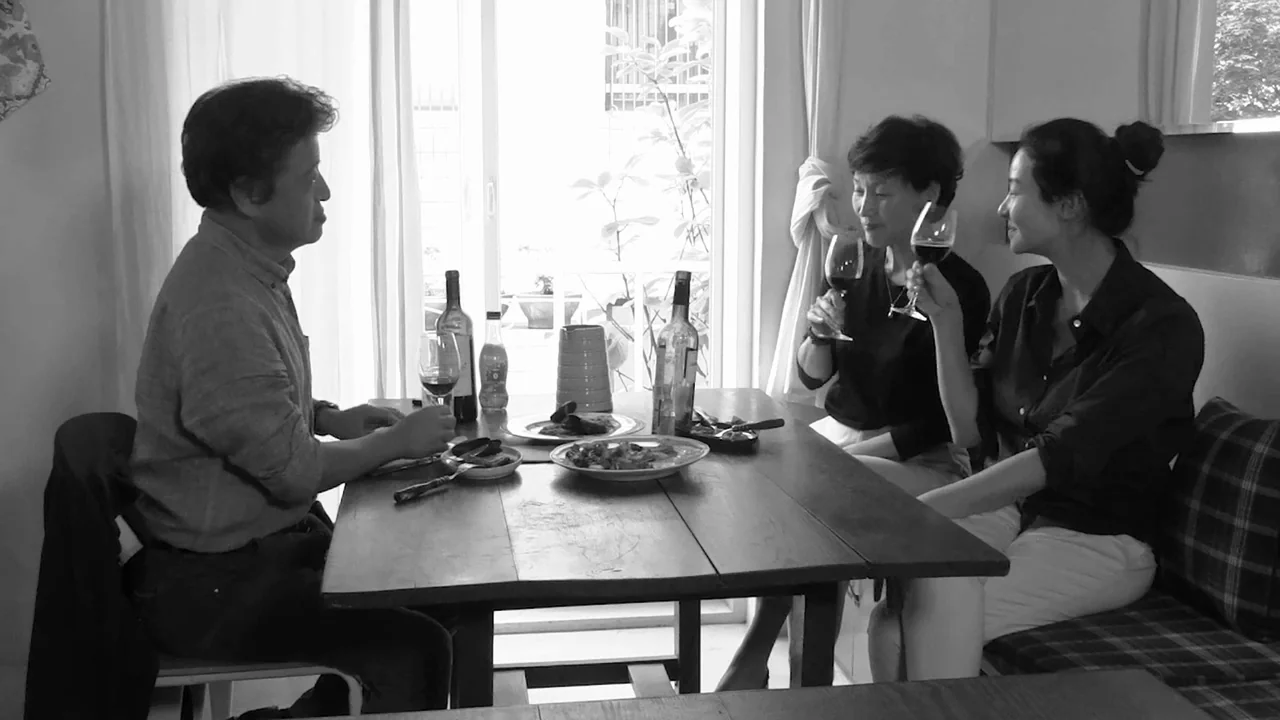 Walk Up (2022) movie still: medium — A group of three people sits around a wooden table in a bright room. The individuals are holding wine glasses…