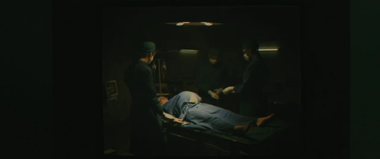 Evolution (2015) movie still: wide — A dark, clinical operating room is illuminated by faint, overhead fluorescent lights. Three surgeons in scrubs…