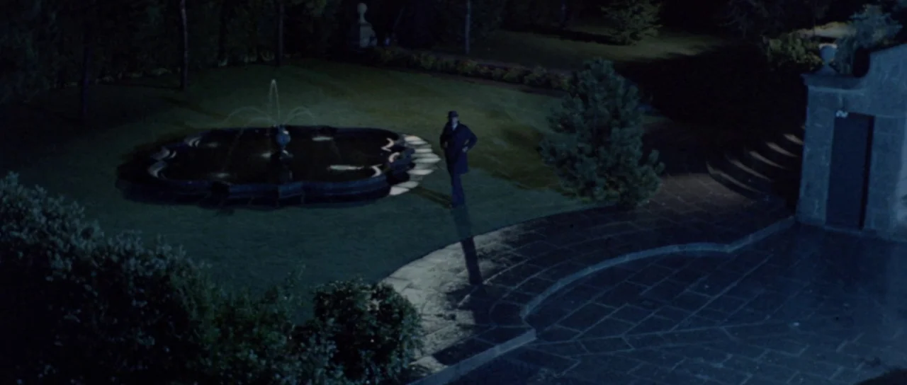 Orgasmo (1969) movie still: wide — A stone courtyard at night features a central fountain spraying water onto the grass. A lone man wearing a coat…