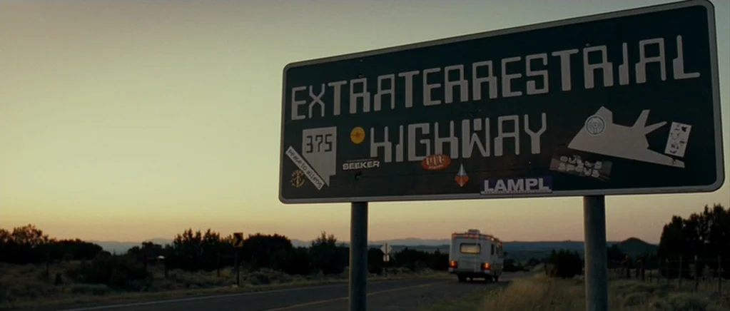 Paul (2011) movie still: wide — A dark green highway sign reading Extraterrestrial Highway stands prominently in the foreground. A camper van drives…