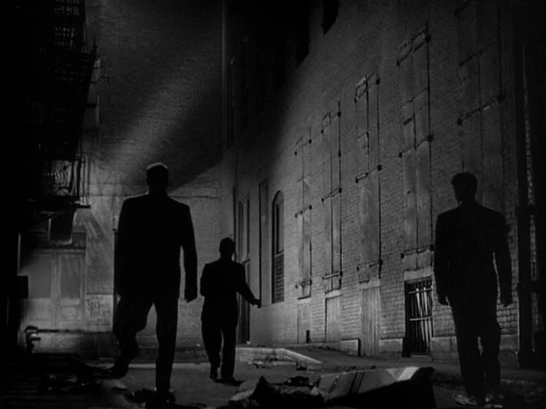 Killer’s Kiss (1955) movie still: wide — A dark brick building with boarded-up windows looms over a narrow, paved passage at night. Three men in…