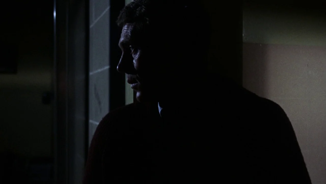 Bullitt (1968) movie still: medium — A dimly lit indoor corridor with plain walls and low illumination. A man stands at the edge of the shadow,…